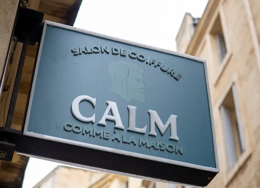 Enseigne suspendue « CALM — salon de coiffure — comme à la maison » dans une rue de Montpellier.